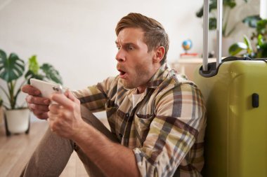 Bavullu bir adam, yerde oturuyor, telefon ekranına bakıyor. Turizm konsepti