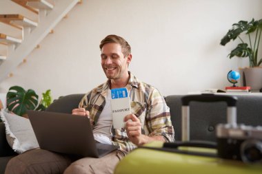 İnternetten uçuşunu kontrol eden, dizüstü bilgisayara bakan, elinde biletlerle evde bavulla oturan neşeli genç adamın portresi..