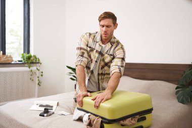 Yatakta oturan bir adamın portresi, valizi kapatmaya çabalayan, tatil için hazırlanan, tatile çıkan. Turizm ve seyahat kavramı