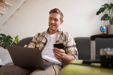 Kredi kartı ve dizüstü bilgisayarıyla gülümseyen yakışıklı adam, tatil için otel rezervasyonu yapan turist, valizin yanında oturuyor..