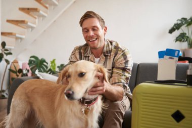 Bavullu, mutlu, yakışıklı genç bir adamın portresi, köpeğiyle oturma odasında oturuyor, tatile çıkmaya hazırlanıyor, tatile çıkıyor. Turizm konsepti