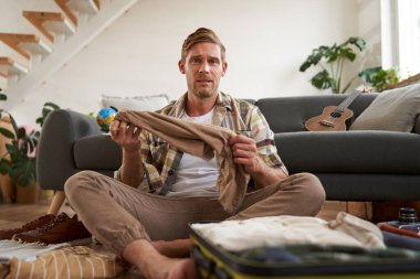 Kafası karışmış bir adamın portresi, elinde bavulla oturma odasında oturuyor, bir pantolon tutuyor, habersiz görünüyor, tatil için toplanmaya çabalıyor. Turizm ve seyahat kavramı