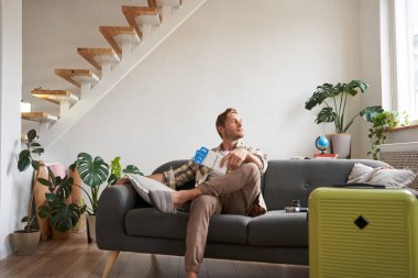 Yakışıklı bir gencin portresi, oturma odasında bavuluyla oturuyor, iki uçak bileti tutuyor, tatile çıkıyor, taksi bekliyor, pencereden dışarı bakıyor. Turizm konsepti