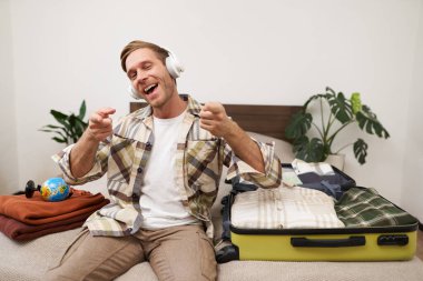 Sarışın, kulaklıklı yakışıklı bir adam, bavul ve kıyafetlerle yatakta oturuyor, tatil için hazırlanırken kablosuz kulaklıkla müzik dinliyor, seyahate çıkıyor..