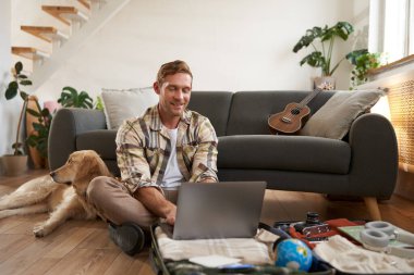 Gülümseyen genç bir adamın portresi, köpekli bir turist, valizini topluyor, tatile çıkıyor, dizüstü bilgisayarına bakıyor, bir otel ya da tatil için uçak bileti ayırtıyor..