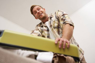 Kaslı, güçlü bir gencin alt açılı portresi. Elbiseleriyle dolu valizini kapatmaya çalışıyor. Çantasına gayretle bastırıyor. Turizm ve yaz tatili konsepti