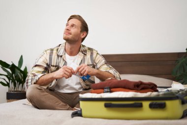 Yakışıklı bir adamın portresi, turist, tatil biletlerine bakıyor seyahat acentesinde yer ayırtıyor, yatakta oturuyor ve giysilerini bavuluna koyuyor..