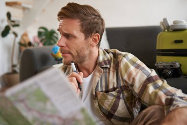 Seyahat haritasına kafası karışmış bir adamın portresi, rotayı seçmek, tatile gitmek, tatile hazırlanmak, oturma odasında bavulla oturmak..