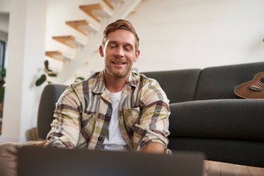 Yakışıklı bir gencin portresi, oturma odasında oturuyor, dizüstü bilgisayar kullanıyor, bilgisayar ekranına bakıyor..