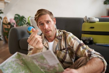 Yol haritası olan yakışıklı bir adam, kalp hareketi gösteriyor, oturma odasında bavuluyla oturuyor, tatil planları yapıyor..