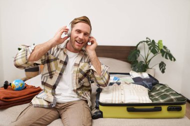 Genç mutlu bir adamın portresi, kulaklıkla müzik dinliyor, otel odasında bavuluyla oturuyor, tatilde giysilerini açıyor, turist tatile çıkıyor. Yaşam tarzı ve insan kavramı