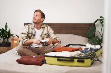 Turist portresi, bavuluyla yatakta oturan, kamera taşıyan, bavulunu kıyafetlerle dolduran, tatile çıkan, yaz tatiline hazırlanan, yurt dışına seyahat eden bir adam..