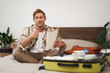 Bavullu bir adamın portresi, tatil için otururken ve eşyalarını toplarken, tatil için eşya listesini kontrol ederken, not alırken, not defterine not alırken. Yaşam tarzı ve turizm konsepti