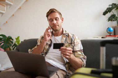 Kredi kartlı genç bir adamın portresi, oturuyor ve dizüstü bilgisayar kullanıyor, bir telefon görüşmesi yapıyor, otel ya da bilet ayırtıyor, telefonla satın alımını onaylıyor, yanında bavulu var..