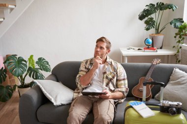 Tabletli genç bir adamın resmi, oturma odasında bavuluyla oturuyor, okuyor, rotayı okuyor, tatile hazırlanıyor. Turizm konsepti