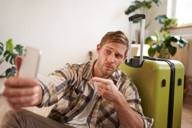Karizmatik maço adam, gezgin akıllı telefondan selfie çekiyor, tatilini videoya kaydediyor, cep telefonu kullanıyor, valizin yanında oturuyor..