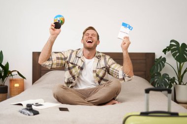 Mutlu adamın portresi, turist yurt dışına tatile çıkıyor, elinde dünya ve uçak bileti var, tatili için heyecanlı, gülümseyen, coşkulu bir yüz ifadesiyle yatakta oturuyor. Boşluğu kopyala