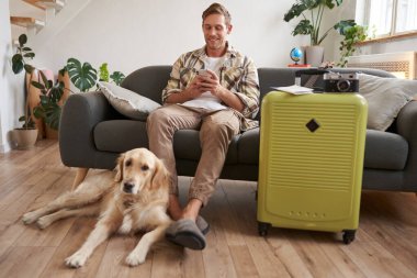 Köpekli bir adamın portresi, bavulunu hazırladı, yurtdışına gitmek için kamera ve fotoğraf çekmek için, köpek bacaklarının yanında dinleniyor, adam cep telefonuna bakıyor ve internetten sohbet ediyor..