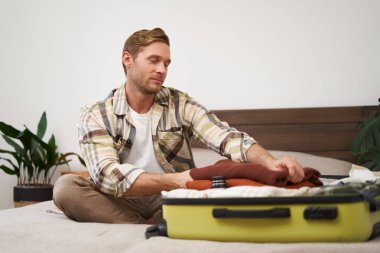 Giysilerini bavula dolduran genç bir adamın portresi, bavulla yatakta oturan, tatile çıkan, bir yolculuğa hazırlanan. Yaşam tarzı ve turizm konsepti