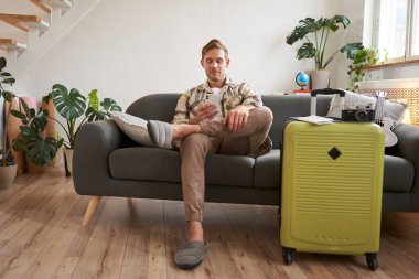 Adamın portresi oturma odasında cep telefonuyla oturuyor, bavulunu hazırlıyor, akıllı telefon uygulamasında sohbet ediyor, tatil yerlerine bakıyor, tatile çıkıyor..