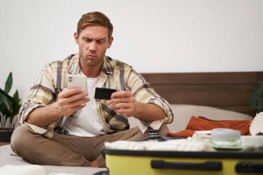 Düşünceli yüzlü bir adamın portresi, bavulla yatakta oturuyor, internetten kredi kartı ve akıllı telefon uygulaması ile ödeme yapıyor. Boşluğu kopyala