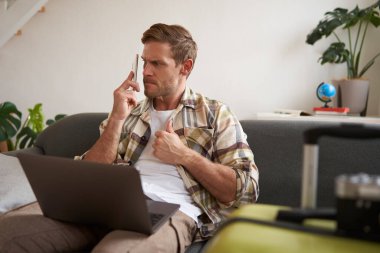 Kızgın bir adamın portresi hatta bekliyor, birini aramaya çalışıyor, akıllı telefon ve laptop kullanıyor, bavulun yanında oturuyor..