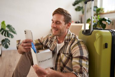 Elinde bavuluyla gülümseyen yakışıklı bir adamın portresi, akıllı telefona bakıyor, seyahat bileti ve pasaport tutuyor. Turizm ve seyahat kavramı