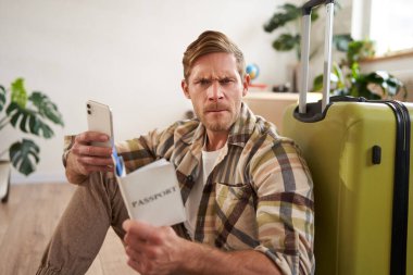 Bavullu ve pasaportlu öfkeli bir adamın portresi, elinde akıllı telefon, kameraya sinirli bakıyor, yerde bavulla oturuyor..