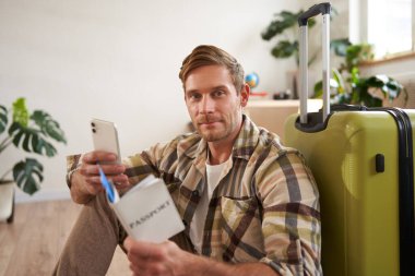 Elinde bavuluyla gülümseyen yakışıklı bir adamın portresi, akıllı telefona bakıyor, seyahat bileti ve pasaport tutuyor. Turizm ve seyahat kavramı