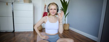 Genç gülümseyen güzel bir kadının portresi, kablosuz kulaklık takıyor, evde çalışıyor, kauçuk yoga minderinde oturuyor, tamam işareti gösteriyor, egzersizlerle tatmin oluyor..