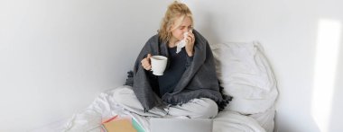 Grip olan, hapşıran, hasta hisseden, dizüstü bilgisayarla yatakta oturup soğuk algınlığı geçiren bir kadının portresi..