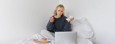 Üzgün, ağlayan genç bir kadının portresi, evde oturuyor, donut ve konforlu yiyeceklerle yatakta oturuyor, dizüstü bilgisayardan üzücü bir şeye bakıyor..
