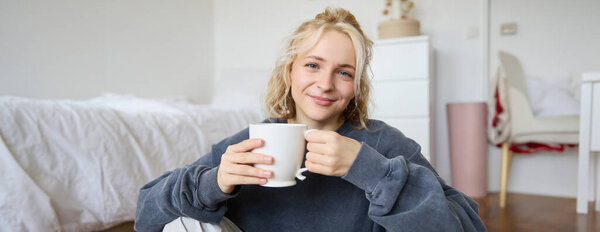Portrait of young woman sitting on bedroom floor, drinking tea, holding white mug and smiling at camera.