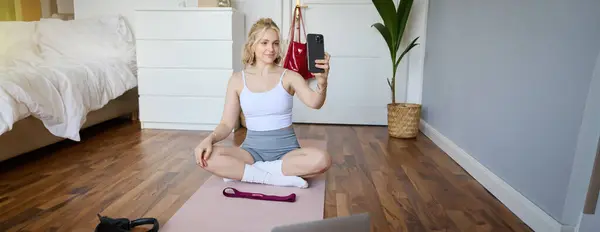 Portrait of stylish young woman, sitting on rubber mat on floor at home, taking selfie while doing workout, yoga session, posting photo on social media.