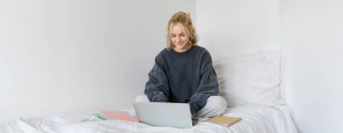 Mutlu genç bir kadın resmi, evden öğrenim görüyor, laptopuyla online kursa bağlanıyor, not defterleriyle yatakta oturuyor, gülümsüyor ve mutlu görünüyor..