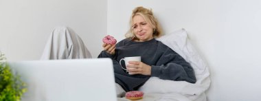 Üzgün, ağlayan genç bir kadının portresi, evde oturuyor, donut ve konforlu yiyeceklerle yatakta oturuyor, dizüstü bilgisayardan üzücü bir şeye bakıyor..
