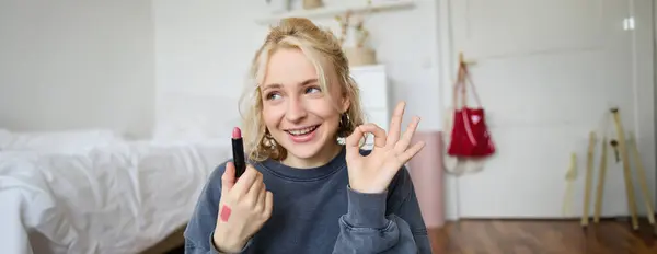 Young female blogger, content creator showing lipstick and okay hand sign, recommending beauty product for her audience on social media, recording vlog in room.