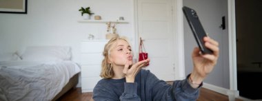 Genç bir kadının resmi yatak odasında yerde oturuyor, akıllı telefondan selfie çekiyor, cep telefonu kamerasına hava öpücüğü gönderiyor, uygulamayı kullanarak görüntülü sohbetler yapıyor..
