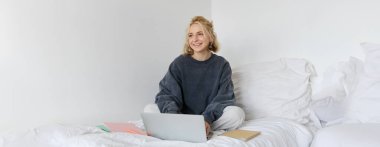 Mutlu genç bir kadın resmi, evden öğrenim görüyor, laptopuyla online kursa bağlanıyor, not defterleriyle yatakta oturuyor, gülümsüyor ve mutlu görünüyor..
