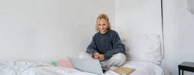 Mutlu sarışın kadın portresi, evde çalışan serbest çalışan, dizüstü bilgisayarları ve defterleriyle yatakta oturan. Yatak odasında ödev yapan öğrenci, görüntülü sohbet yoluyla çevrimiçi sınıfa bağlanıyor.