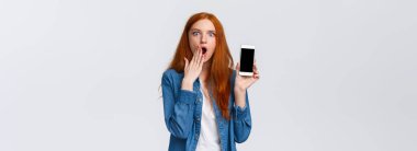 Etkileyici ve şaşkın kızıl saçlı bir kadın inanılmaz bir şey görüyor, internete çay döküyor, nefes nefese kapağını açıyor ve Samrtphone ekranını, beyaz arka planı gösteriyor..