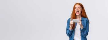 Kaygısız, güzel, bin yıllık kızıl saçlı kız eğleniyor, favori video blogcusunu izliyor, internetteki komik videoya gülüyor, akıllı telefonu yatay olarak tutuyor, beyaz arka planda neşeli bir şekilde duruyor..