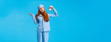 Alarmı neden kurmadın? Kafası karışık ve kararsız, pijama ve uyku maskeli çekici kızıl saçlı, kafası karışmış, sorgulanmış kameraya bakıyor, pelerin giyiyor, mavi arka plan.
