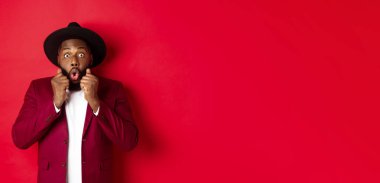 Moda ve parti konsepti. Şaşırmış afro-amerikan erkek model şaşkın şaşkın bakıyor ve hayret verici ve dik dik bakan kamera diyor, kırmızı arka planda duruyor.