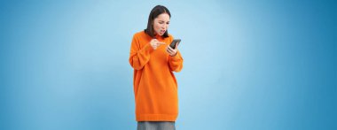 Genç Asyalı kadın cep telefonundan saldırgan yorumlar okuyor, kameraya bakıyor ve hayal kırıklığına uğruyor, akıllı telefonuyla aşağılanıyor..