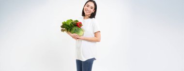 Gülümseyen Asyalı kadın, organik yeşil sebzeleri gösteriyor, sağlıklı çiğ yiyecekler yiyor, beyaz arka plan.