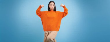 Güçlü ve sağlıklı insanlar. Genç Asyalı kadın kaslarını gösteriyor, kaslarını kasıyor ve kendinden emin görünüyor, gülümsüyor. Keyfi yerinde, mavi arka planda..