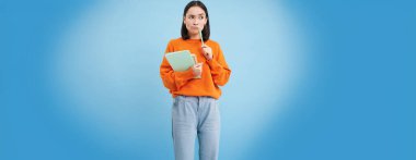 Asyalı bir kız, düşünen bir öğrenci, ciddi bir yüzle smth düşünüyor, elinde kalem ve defterler, mavi arka planda duruyor..