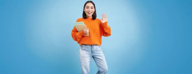 Dost canlısı genç bir bayan, öğrenci merhaba diyor, elinde defterler ve el sallayarak seni selamlıyor, mavi arka planda duruyor..