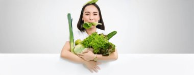 Gülümseyen Koreli kadının portresi marul yaprağı yiyor, elinde yeşil sebzeler, besleyici yiyecekler, beyaz arka plan.